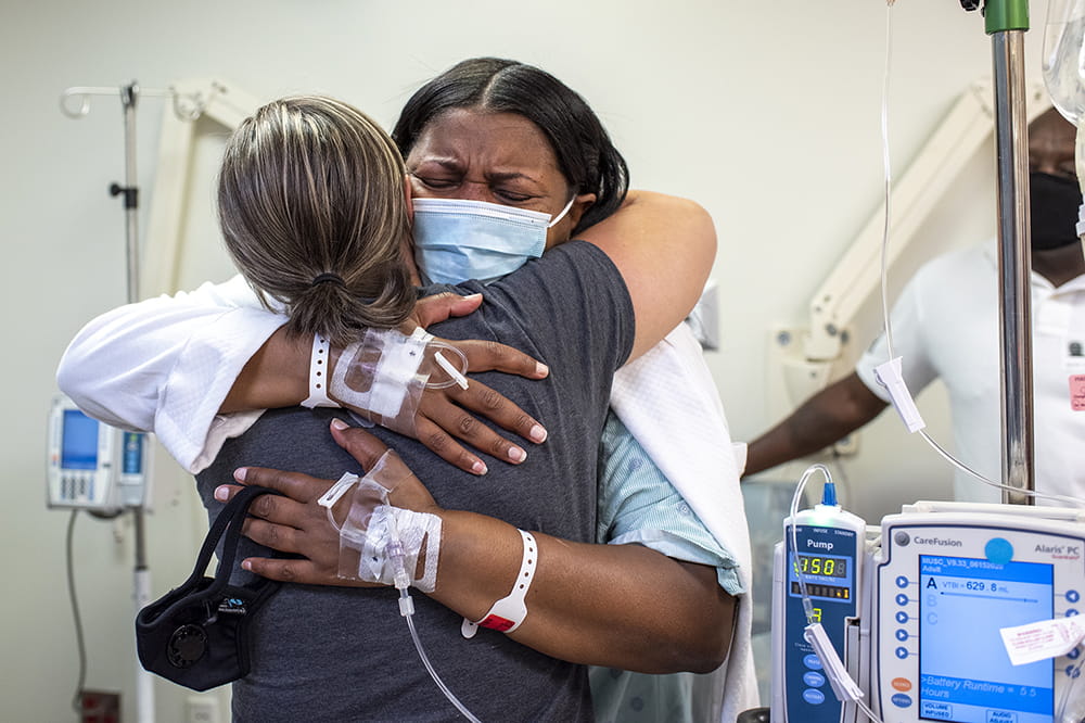 a woman in a hospital gown with IVs in her hands cries and hugs another woman