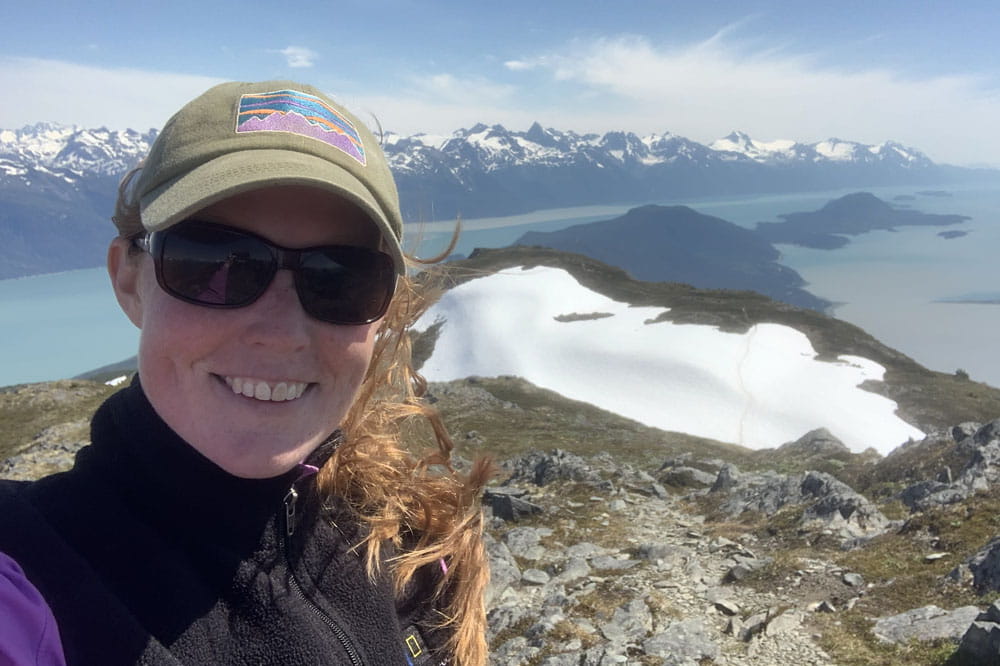 Lucy Boyce hiking in Alaska with snow covered mountains and water in the background