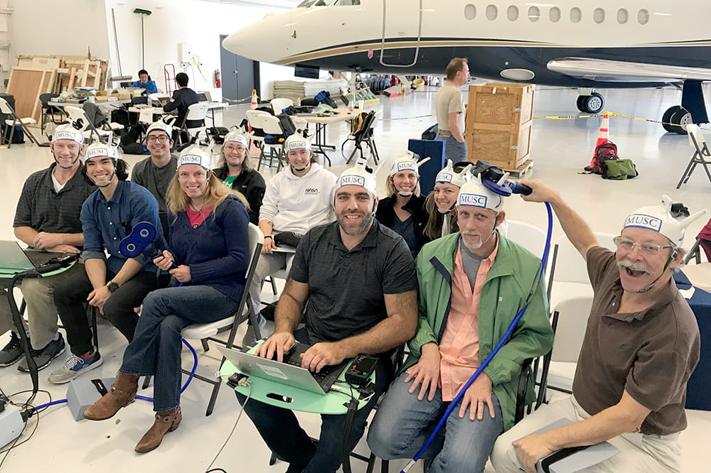 the team sits in an airplane hangar, with a private plane in the background, with the handcrafted helmets on their heads