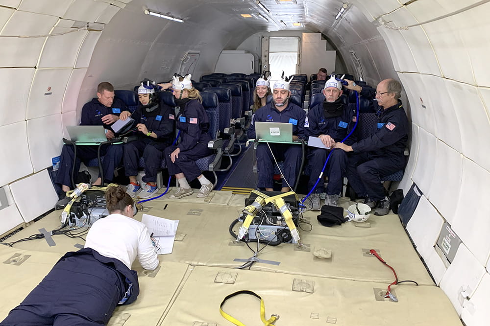 the team sits in seats inside an otherwise empty plane with stark white walls. One team member lays on her stomach in front of the group looking at a laptop