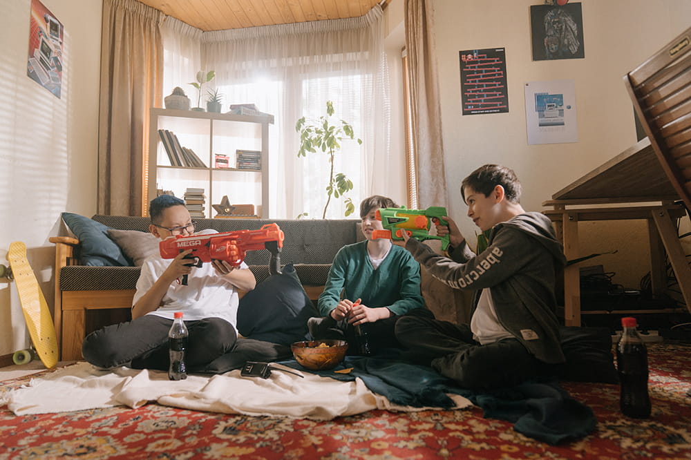 three tween boys sit on the floor of a family room, with two boys playfully pointing toy water guns at each other