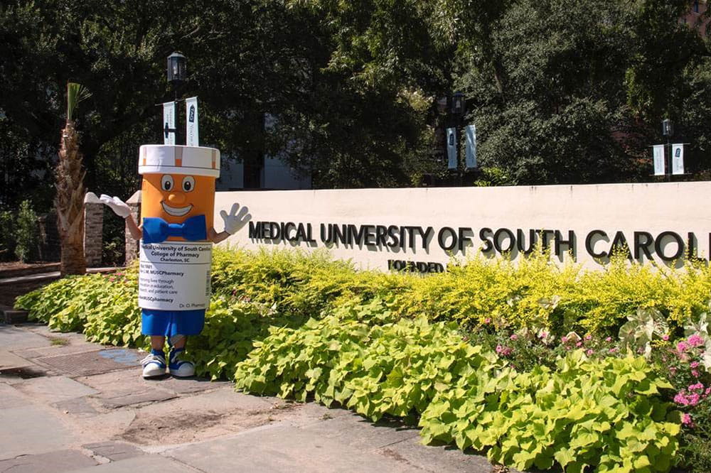 college of pharmacy's pill bottle-shaped mascot stands outside and waves next to MUSC sign