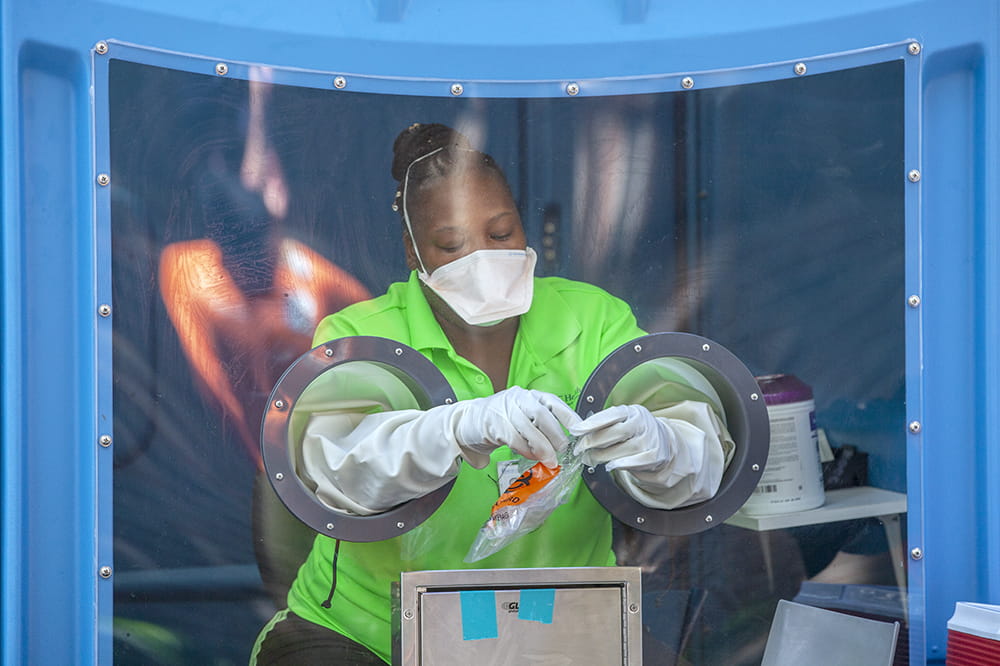 a woman sits inside the blue shell of a portable toilet, wearing a mask over her nose and mouth, reaching her arms through built-in gloves to hold a specimen collection bag