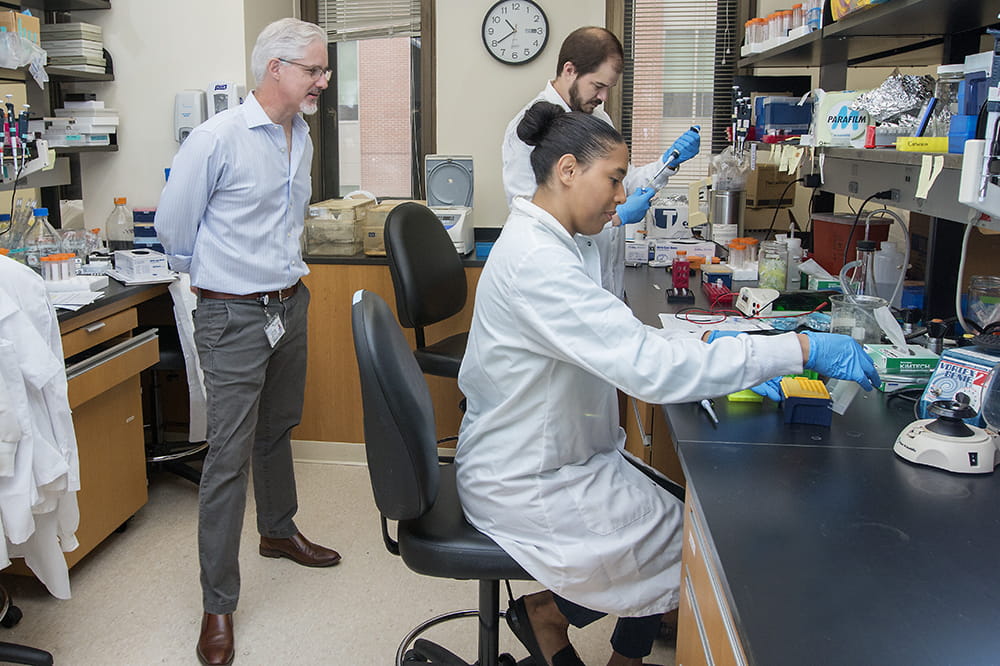 A man and woman sit at a bench in a lab working while their mentor stand back and watches