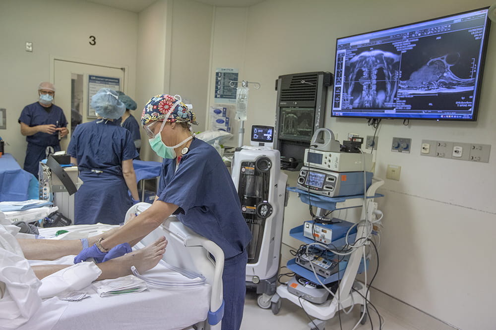 a woman leans over a patient's legs. on the wall behind her is a large screen with an X-ray visible