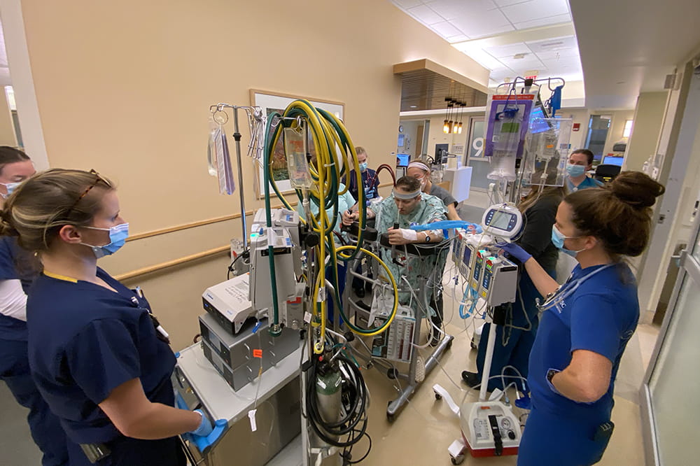 a man walks down to a hospital hallway hooked up to a giant machine while a team of people surround him monitoring his numbers