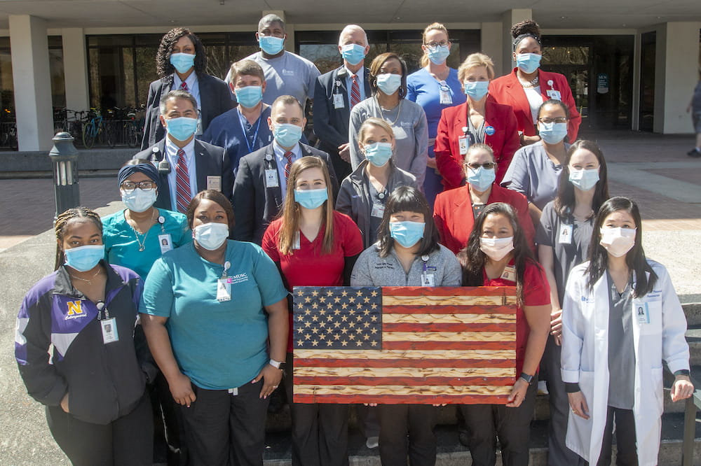 A group of about 20 health care employees standing outside behind a hand carved wooden American flag