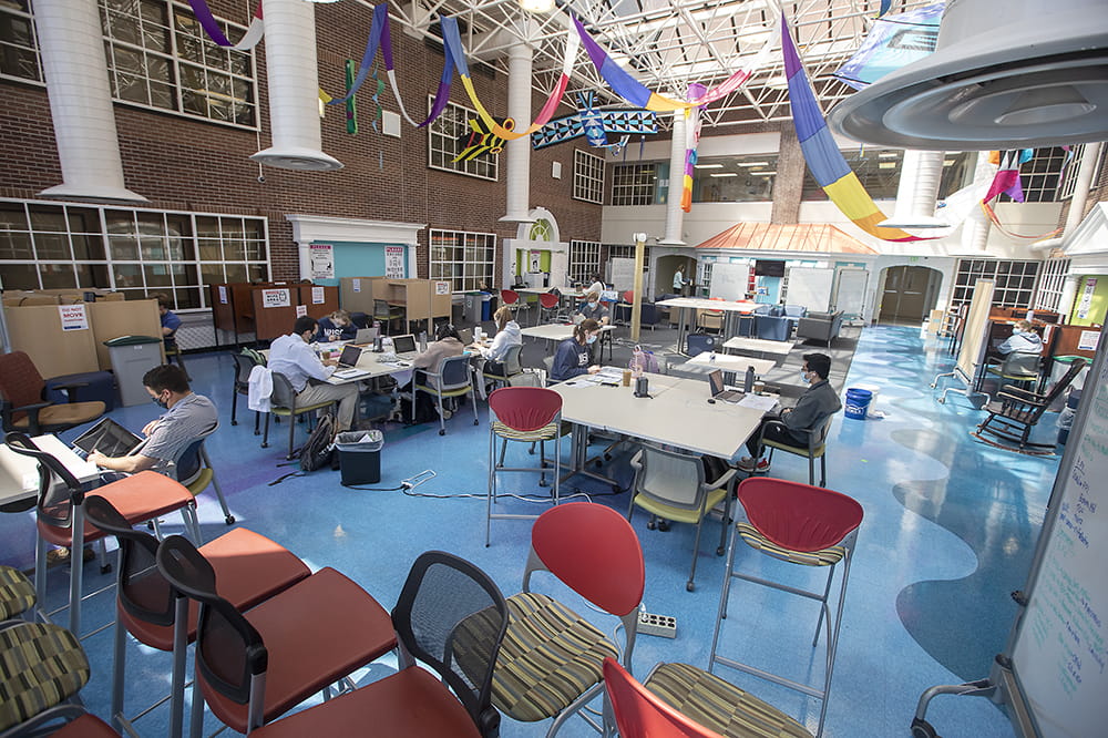 students sit at clusters of tables in a large room, chairs placed haphazardly about the perimeter while colorful streamers still hang from the two story ceiling