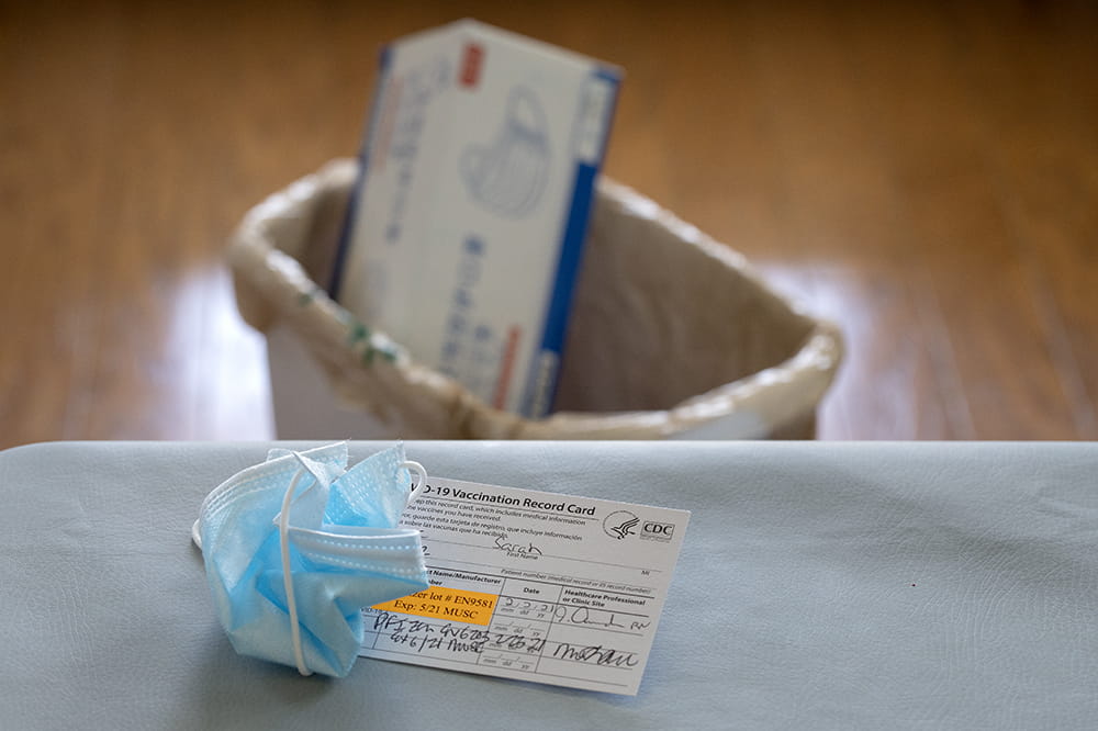 Photo illustration shows a crumpled mask on top of a vaccination card and a box of masks in a trash can.