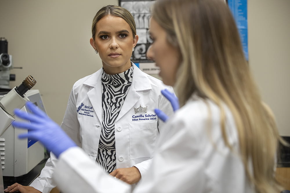 Camille Schrier listens as she wears a specially made Norris Lab lab coat that has her name and Miss America 2020 embroidered on it