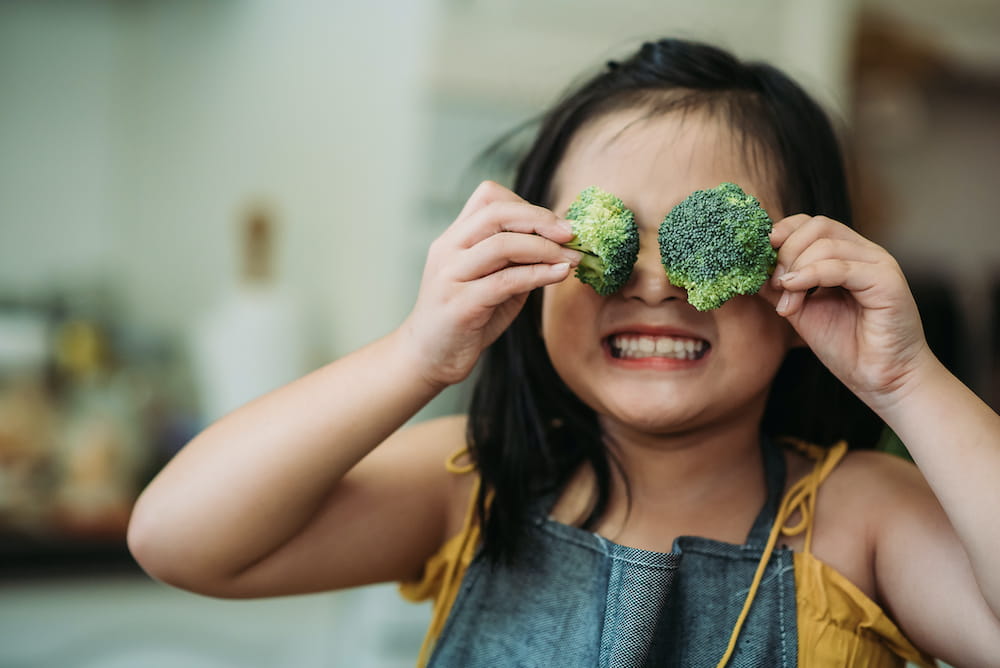 A little girl holding two pieces of raw broccoli in front of her eyes being silly