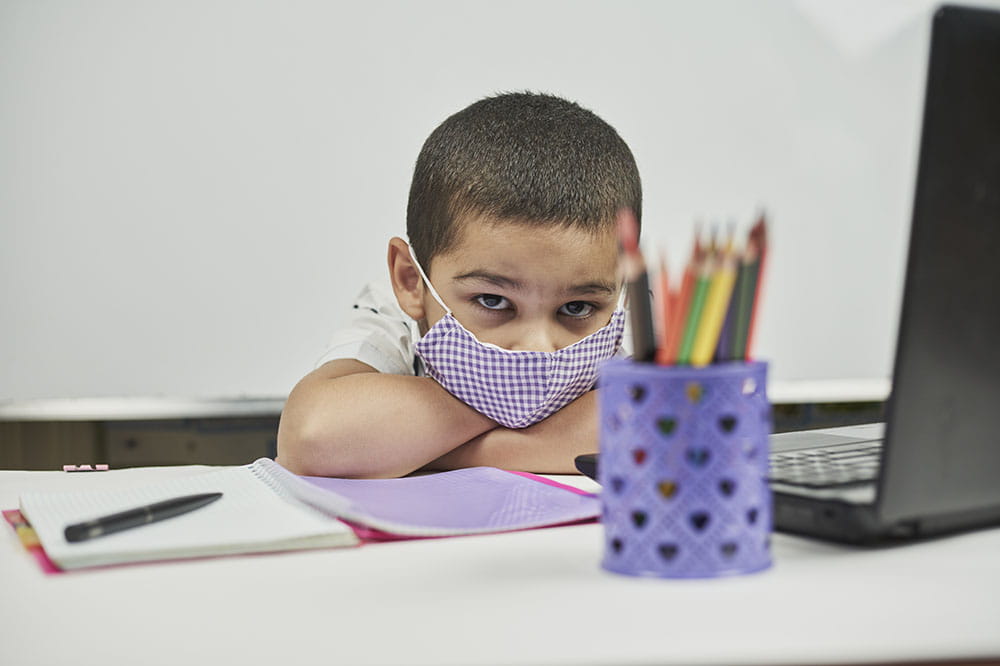 Elementary school age boy looking bored wearing a purple mask and siting by a laptop computer.