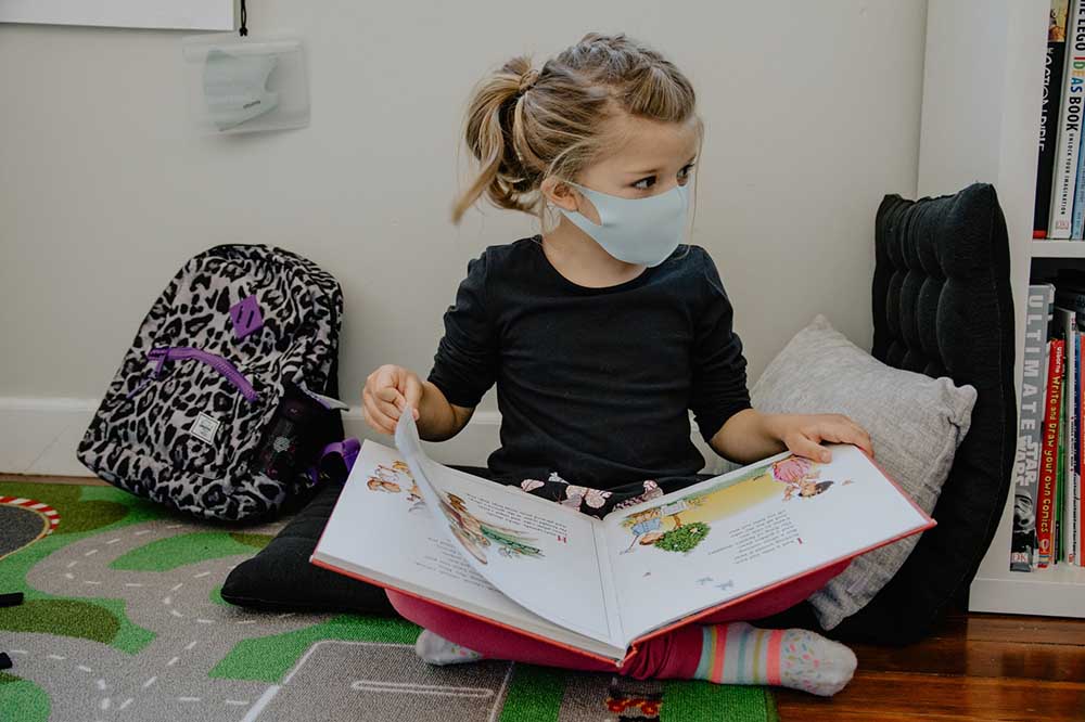 a young girl sits cross legged with a picture book open on her lap while wearing a mask