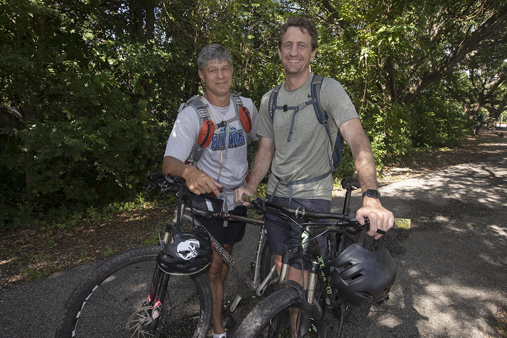 two men pose next to their bikes on a paved trail