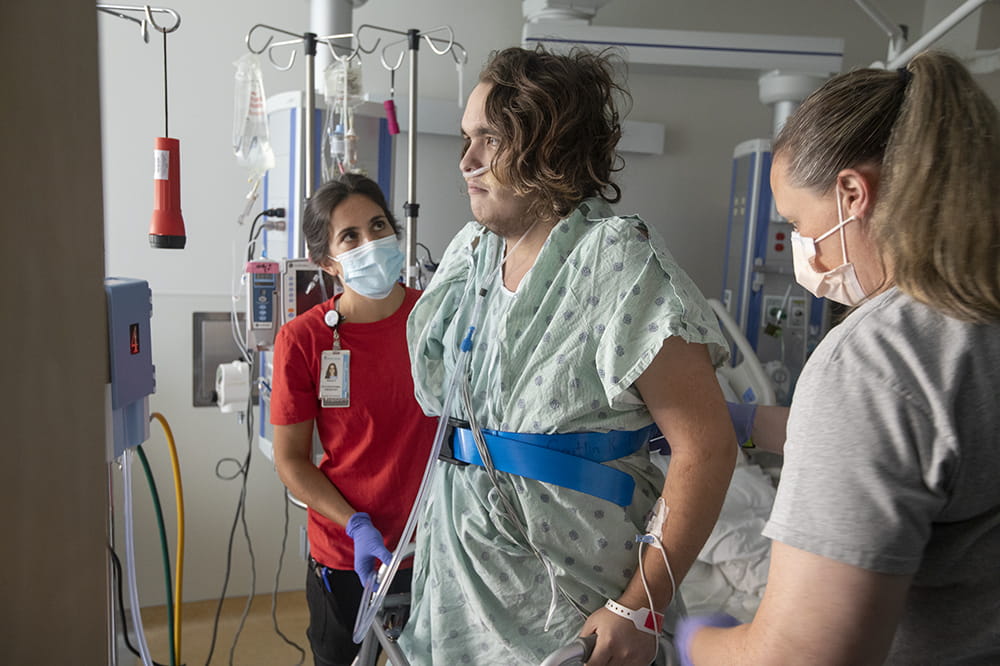 a young man in a hospital gown stands with the help of a walker and two staffers