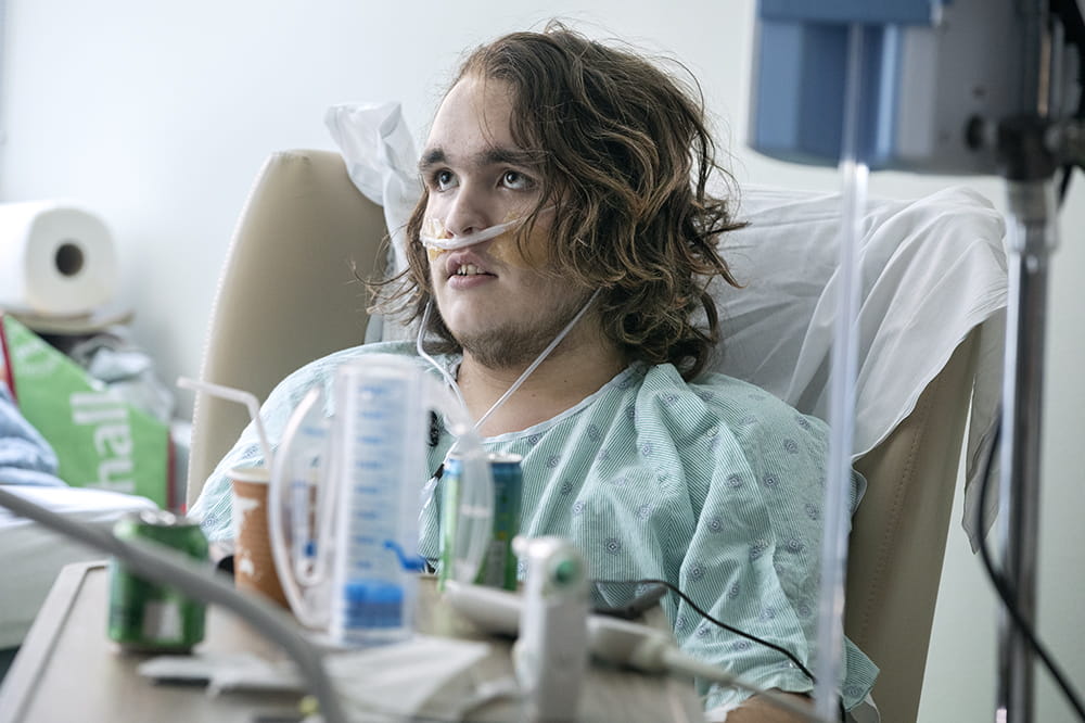 a young man sits in a hospital gown with an oxygen tube attached to his nose