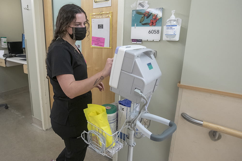 a young woman in black scrubs checks on a patient monitoring machine in a hospital hallway