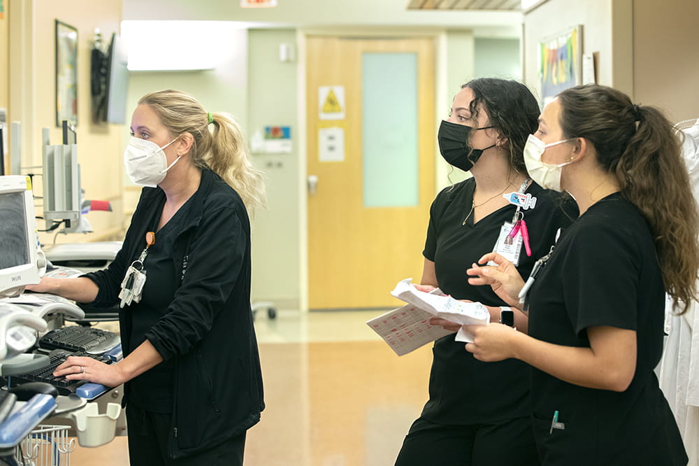 two young women in black scrubs listen to a third woman as she looks over a computer