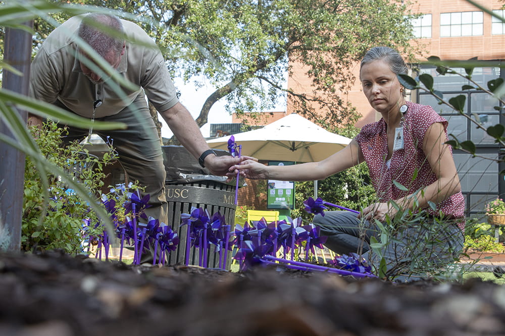 a man and woman place purple pinwheels in a flower bed area