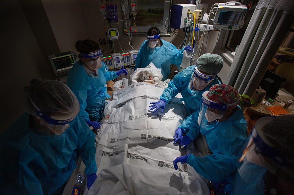 A team of six nurses surround a COVID patient who lies face down on a bed, covered in drypads
