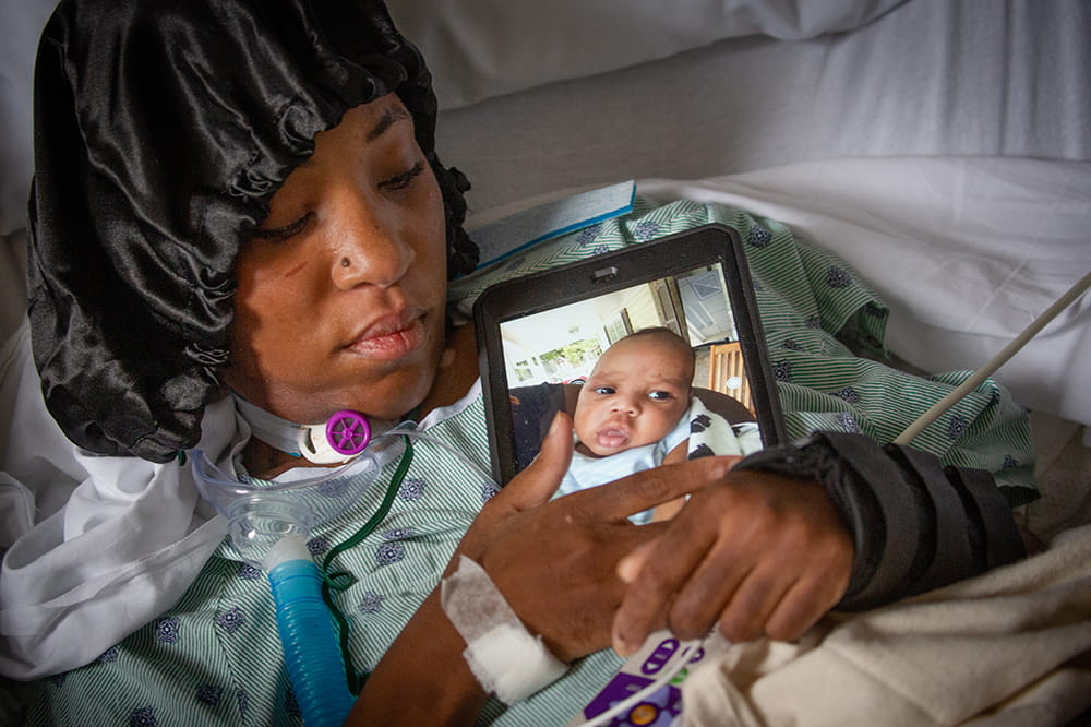 A COVID-positive woman lies in her hospital bed while clutching an iPad showing her little baby boy