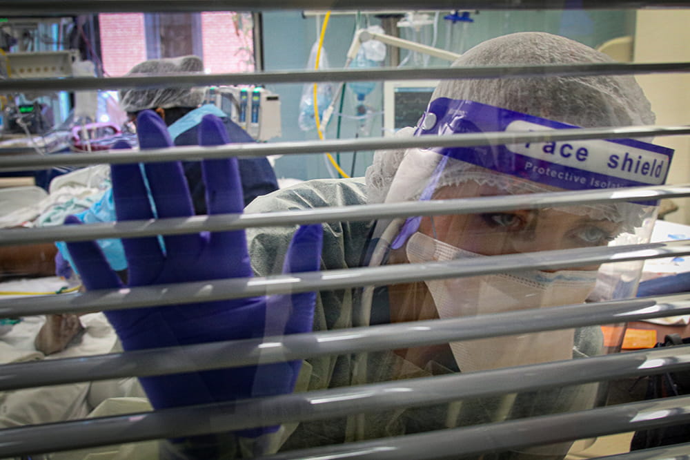 A nurse in full PPE peeks out the window of a isolation room containing a COVID positive patient
