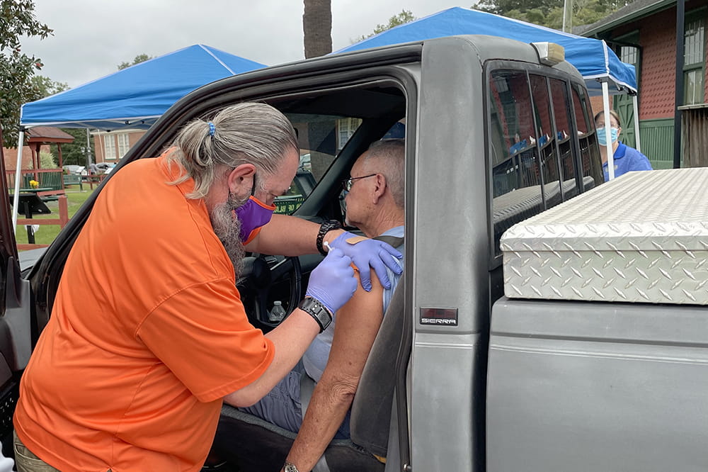 a man in a Clemson orange shirt and purple mask administers a vaccine to a man sitting in the drivers seat of a pickup truck