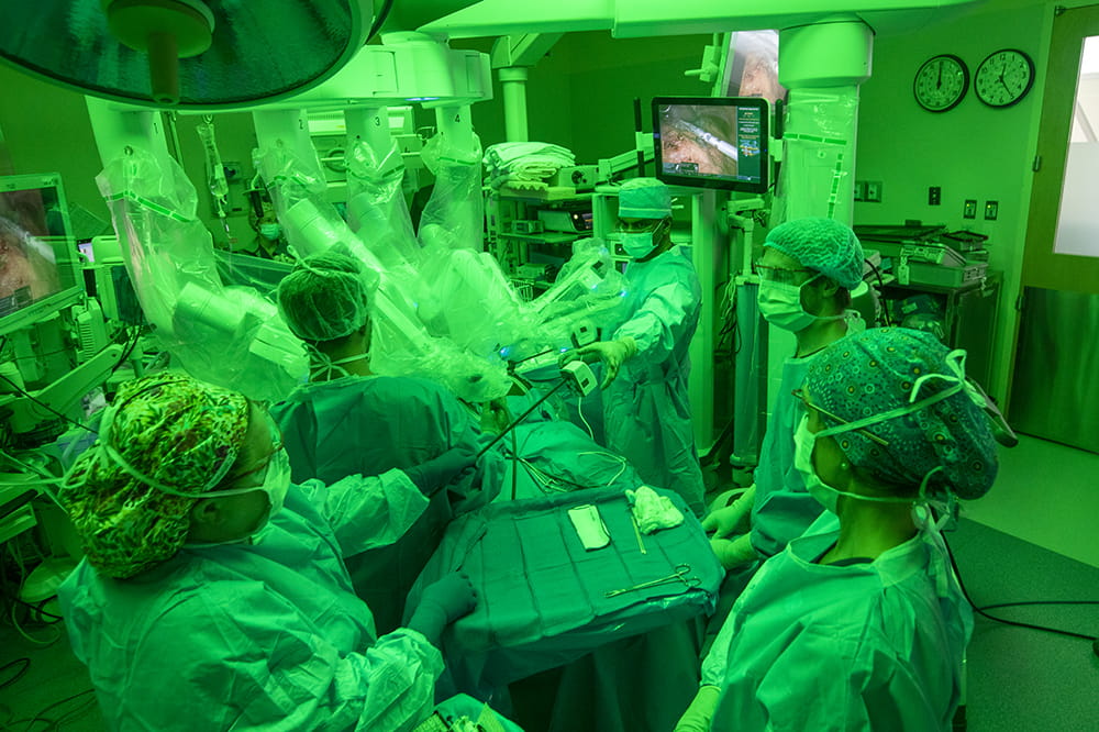 Surgeon Rana Pullatt works in the operating room while surrounded by several other doctors and nurses. They are all bathed in a green light