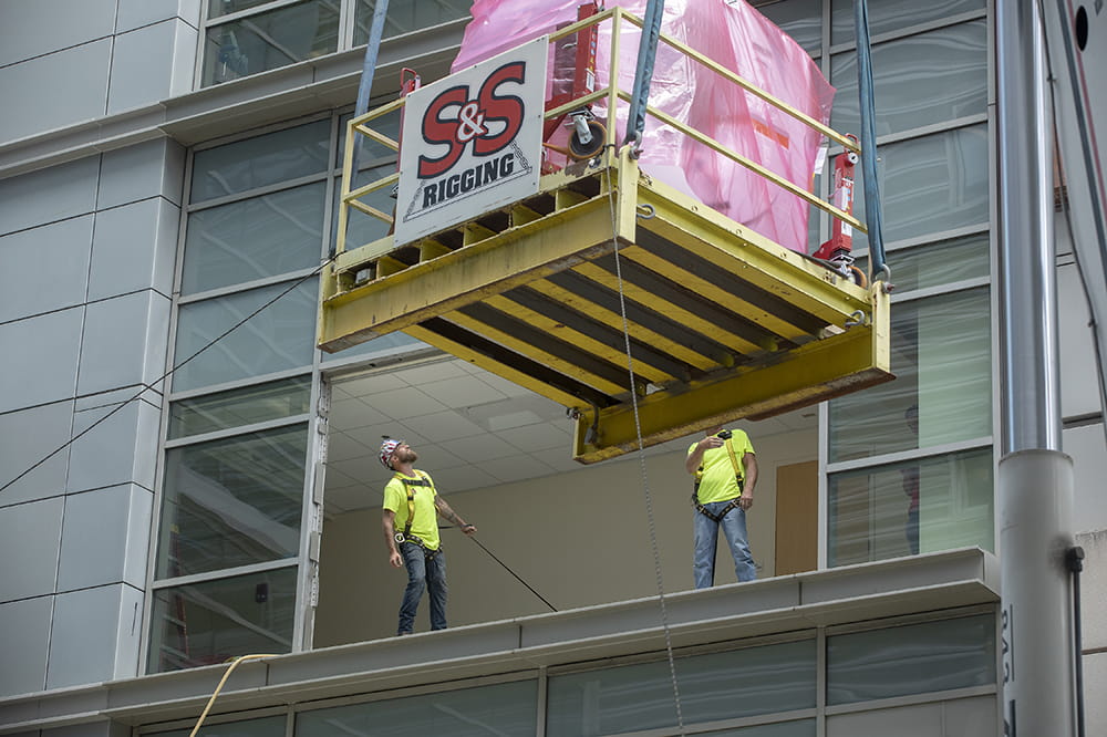 two men standing in an open window are dwarfed by the CT machine, enveloped in pink padding and sitting on a wooden crate, as it is lifted by crane toward the building