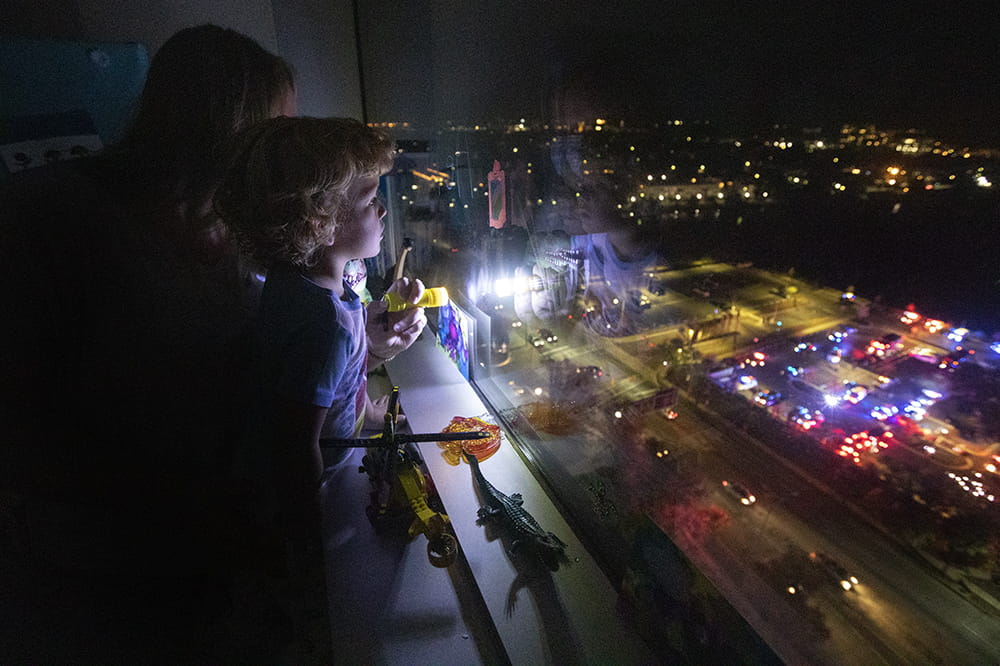 a young boy stands in front of a window in a darkened room with his mom, looking down on lit up first responder vehicles