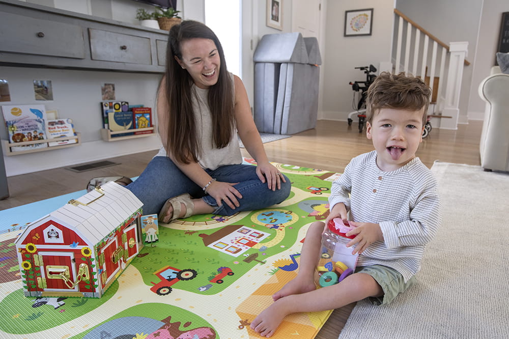 in a bright airy living room with a colorful rug of a farm a little boy grins at the camera while his mother sits a foot or so behind and smiles at him