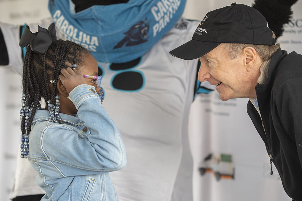 a little girl with beads and bows in her hair puts on a pair of glasses, looking at a smiling man who crouches down to be at eye level; behind them Sir Purr, the Carolina Panthers mascot, looms 