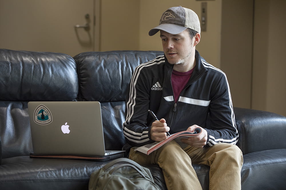 A man wearing a hat sitting on a couch with a book on his lap looking at an open laptop