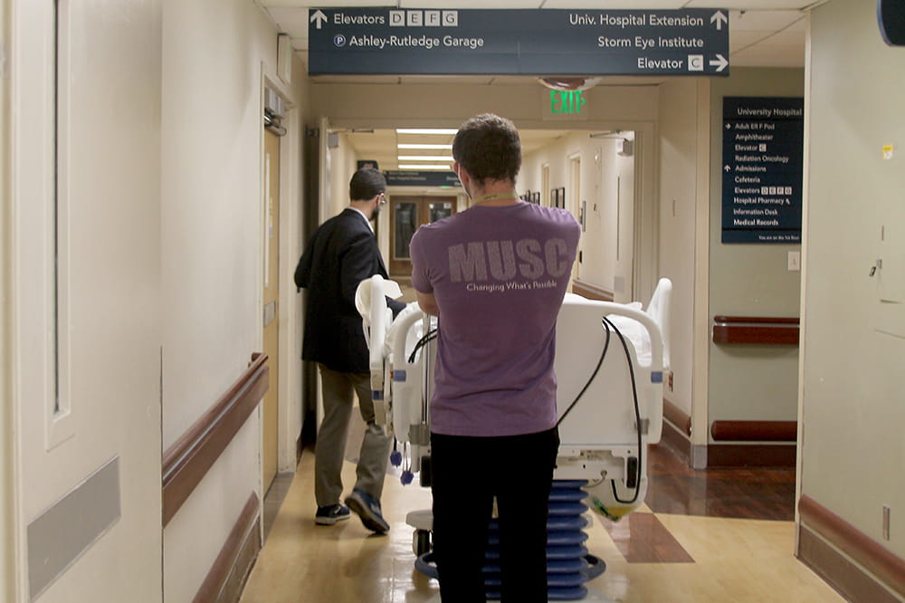 two men, one in office clothes, one in company tee shirt, maneuver a hospital bed through a hallway