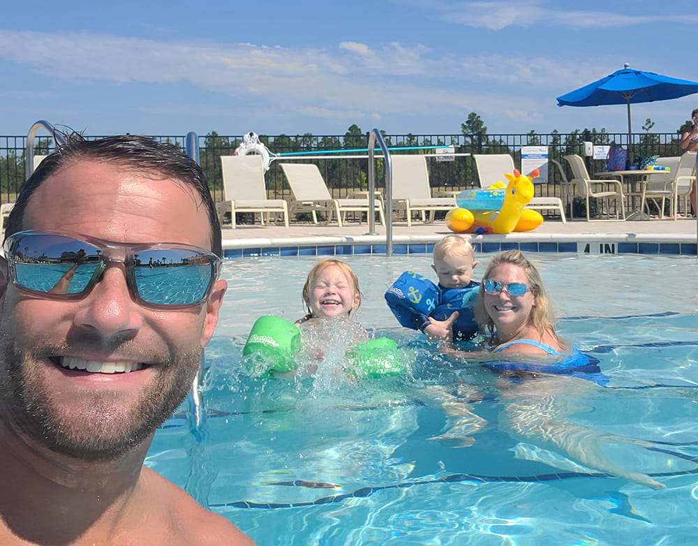 a man smiles into the camera as he takes a selfie in a pool of himself and his smiling family behind him on a nearly cloudless day