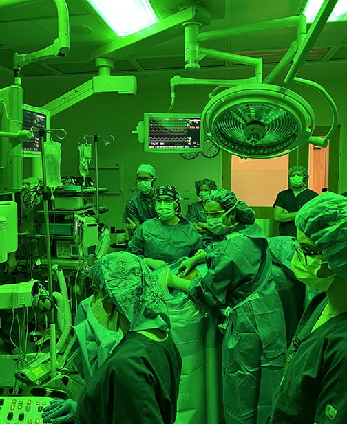 a surgical room is bathed in fluorescent green light as everyone in the room looks at a screen just out of camera range