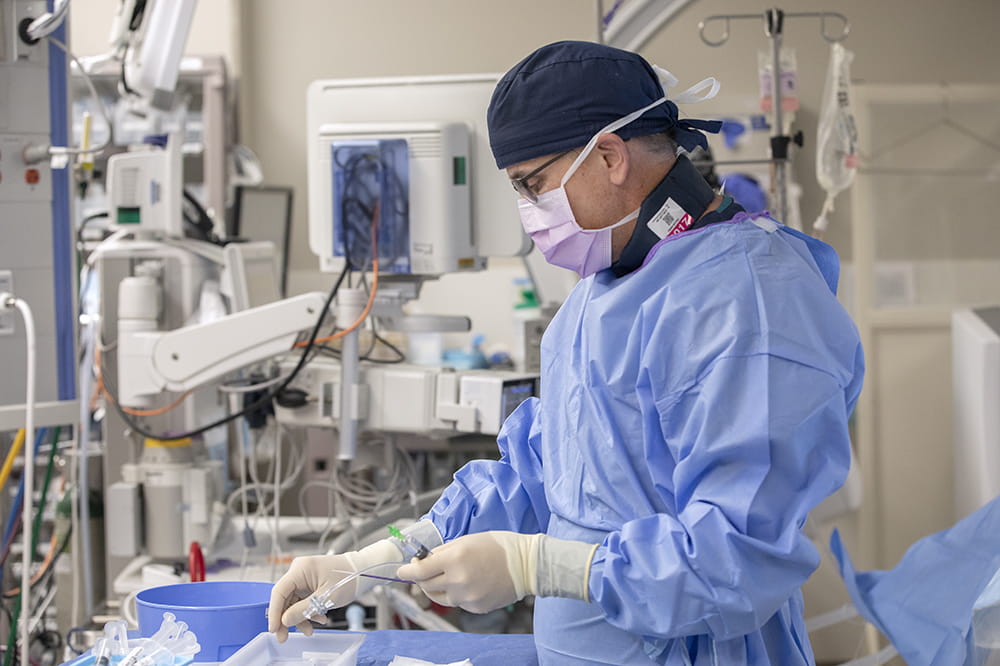a man in surgical cap, gown, gloves and mask prepares instruments in a procedure room