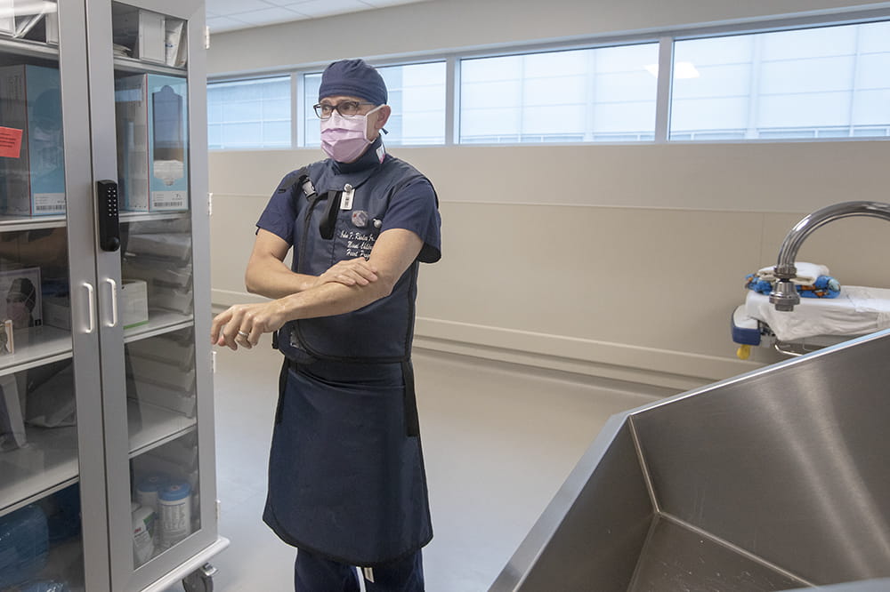 a man in a scrub cap, surgical mask and lead apron washes his arms with soap