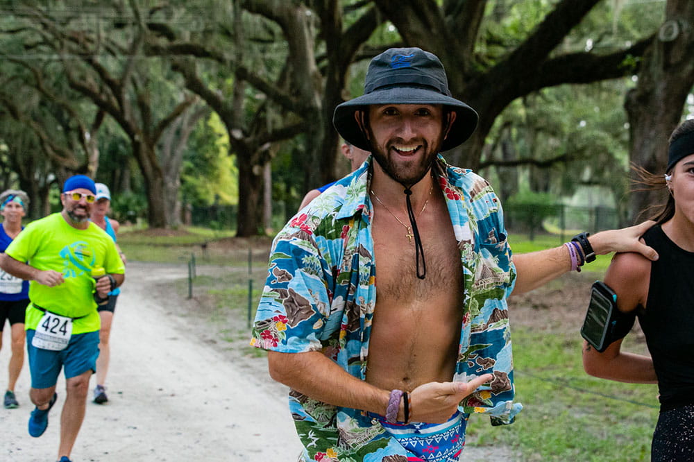 a man running in a race in a tree lined park wears an unbuttoned Hawaiian shirt and floppy hat and grins for the camera while gesturing to the runner next to him