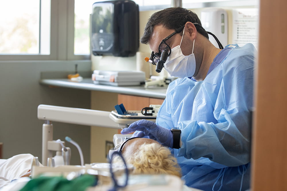 Dental student Keith Tormey examines a patient.  He is masked and wears glasses.  The patient is lying down.  You can see her curly blonde hair.