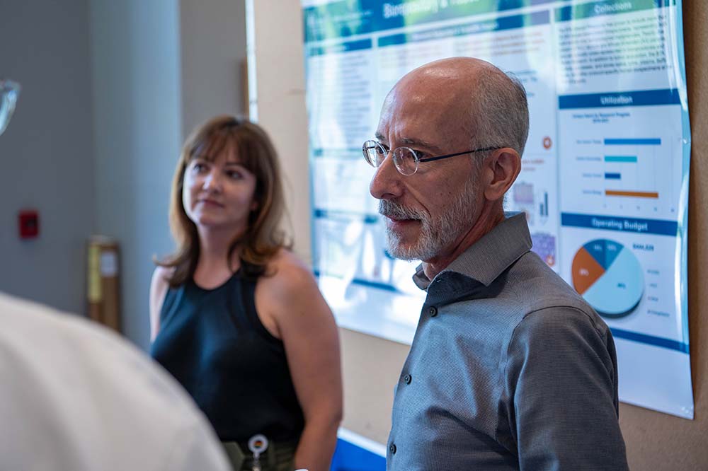 Man wearing glasses speaks while standing in front of a poster. A woman stands to his right listening.