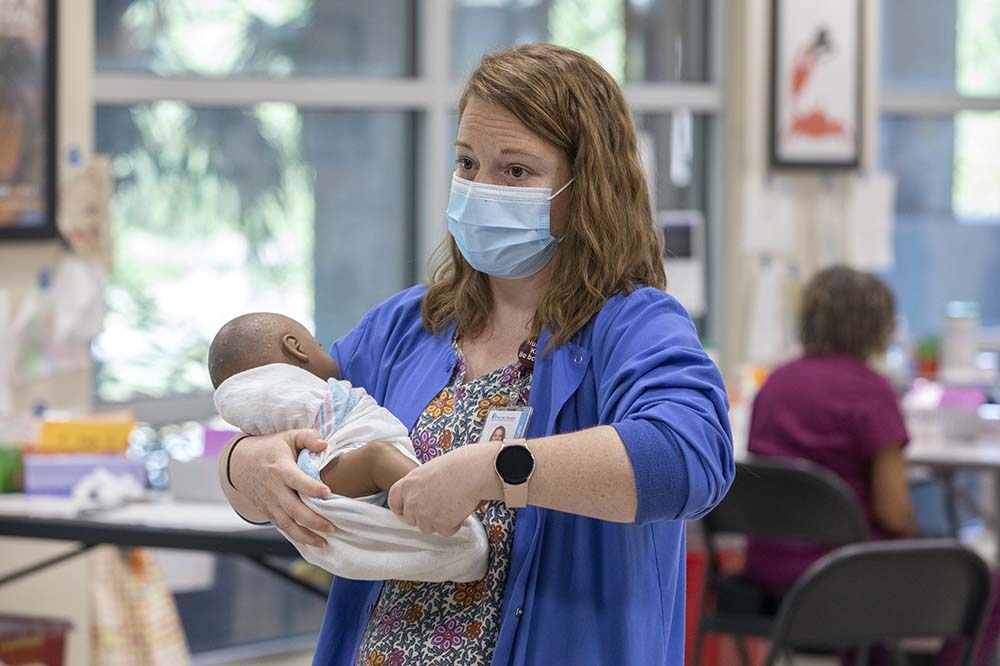 Nurse Haley Jagarwal holds a rubber demo baby. She is wearing a surgical mask and a blue coat. The faux baby is wrapped in a blanket.