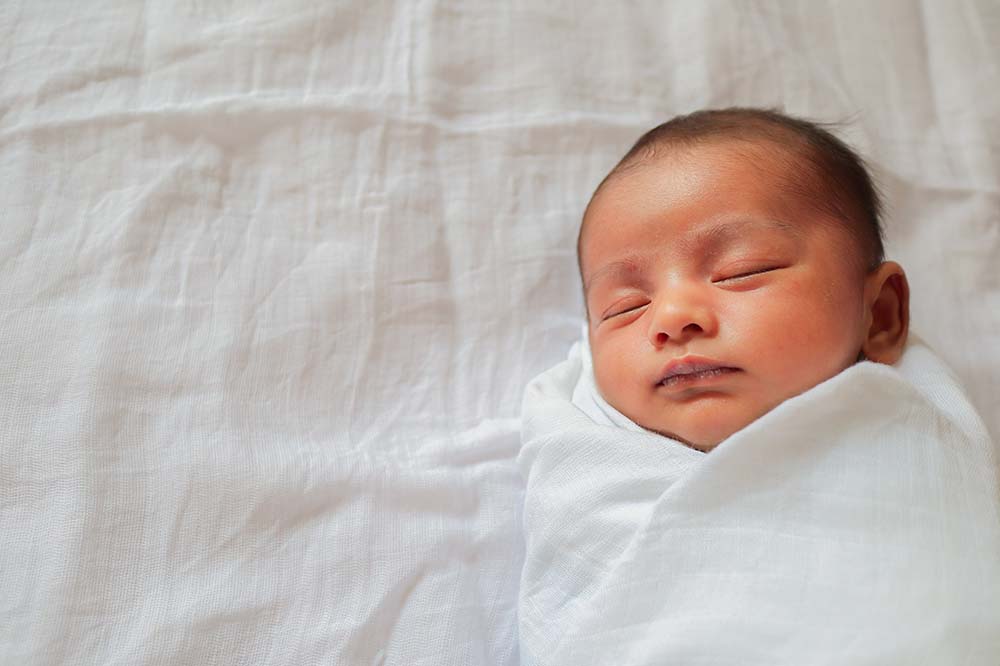 Baby sleeping covered by white wrap lying on white sheet.