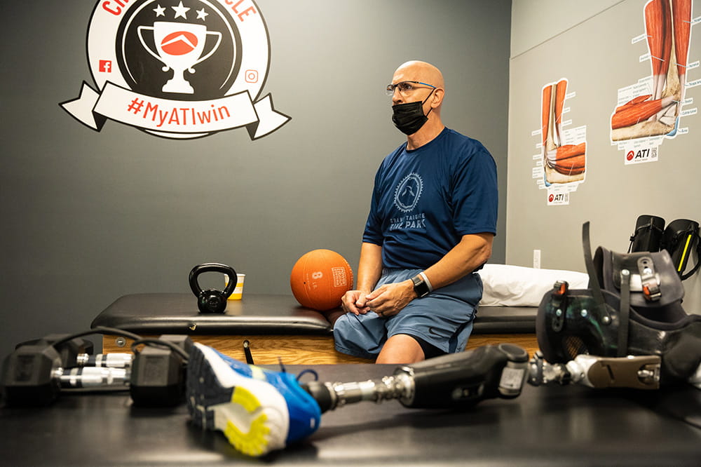 a man in workout gear sits in a rehab room with anatomical pictures of legs on the wall and a prosthetic leg in a running shoe on the table in front of him