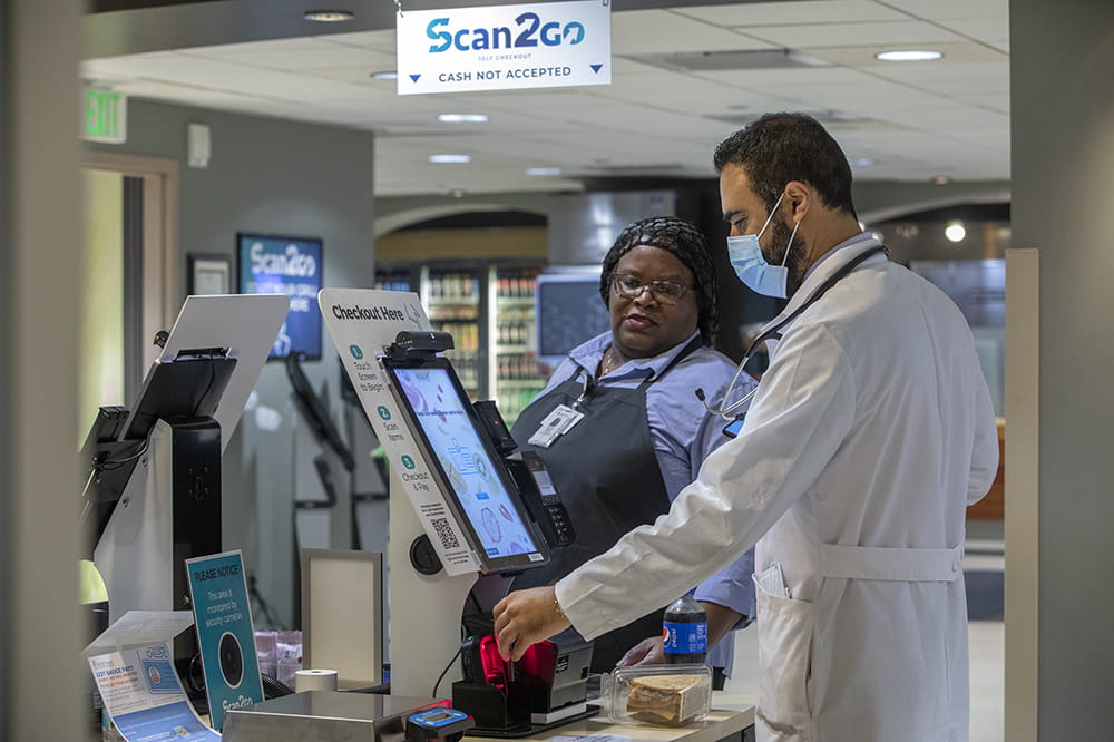 A man in a white doctor's coat looks at a self service checkout while a woman wearing a work apron watches. They are Karim Soliman and Charleen Watson.