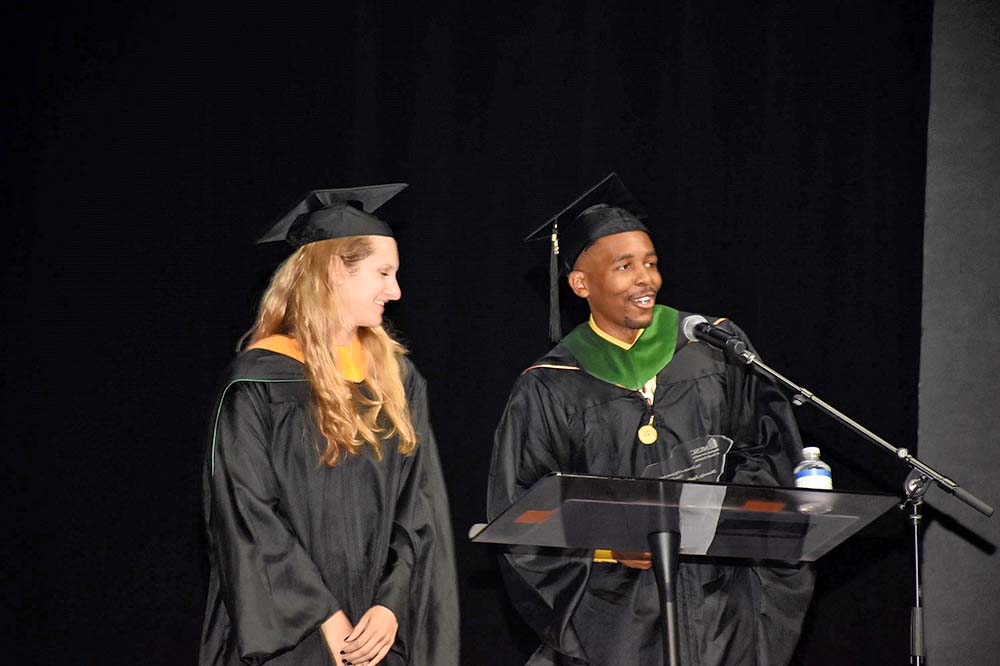 Man in graduation regalia speaks at podium while a woman also in regalia listens. They are wearing black robes and mortarboards.