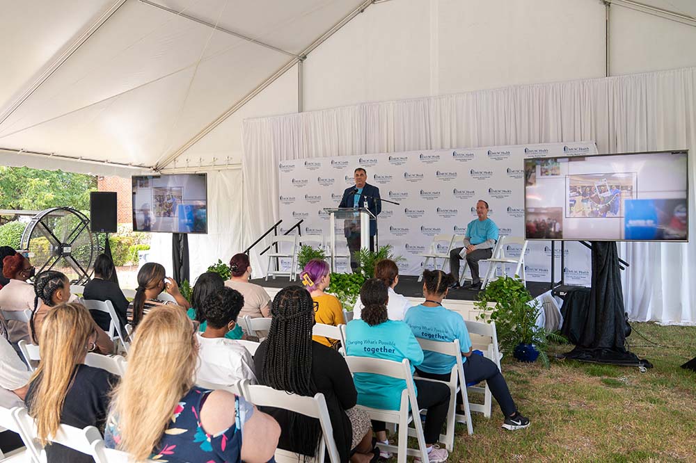 The man at the podium speaks to the group of people sitting on chairs under a canopy.  The speaker is Terry Gunn, CEO of the MUSC Health Midlands Division.
