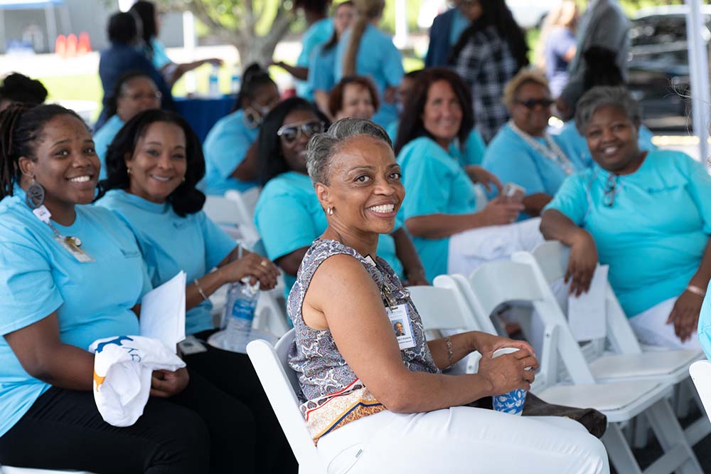 The women sitting on the chairs smile.  One in particular smiles at the camera.  Most are wearing matching blue shirts.