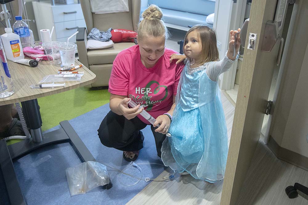 Erika Mann helps with her daughter's medical tube.  Natalyn, 2 years old, wears a blue princess dress.