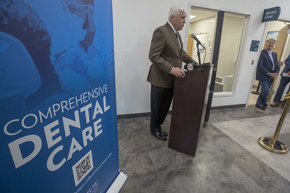 Dr.  David Cole, dressed in a suit, stands at a podium next to a sign advertising dental care.